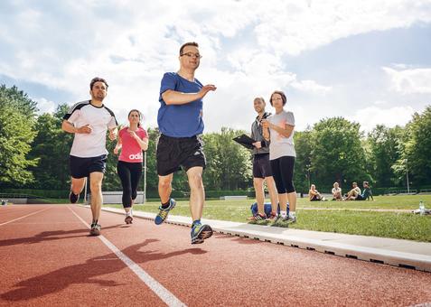Inklusive Laufgruppe mit Läufer mit Prothese auf der Bahn; zwei Personen beobachten mit Stoppuhr und Klemmbrett.