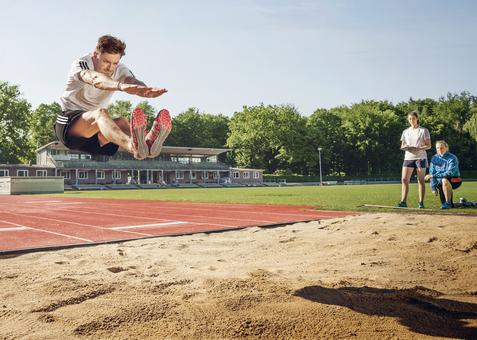 Weitspringer in der Luft beim Absprung über eine Sandgrube, daneben zwei Beobachterinnen.