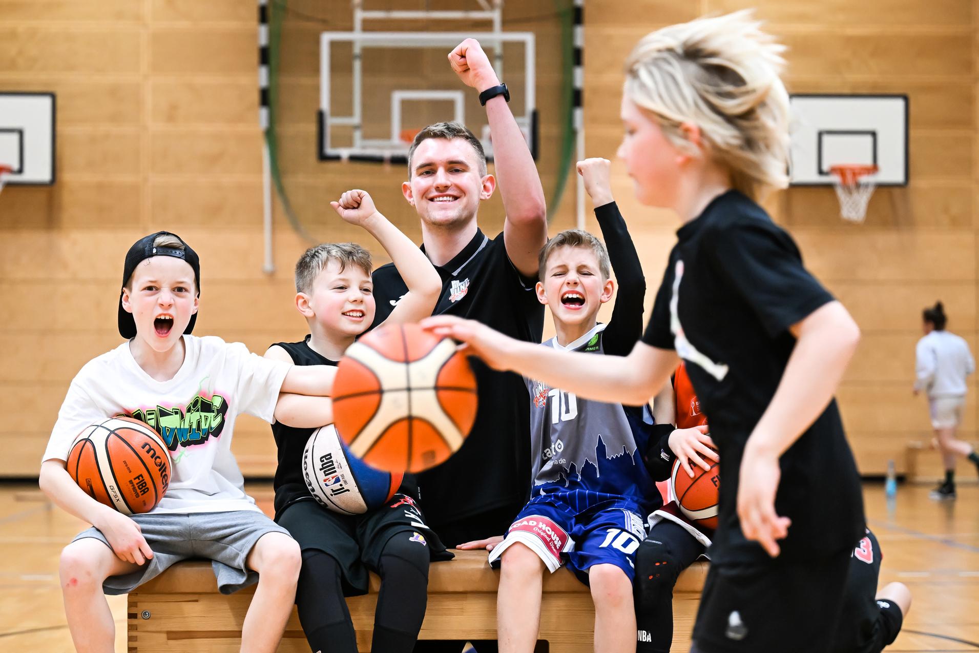 Gruppe fröhlicher Kinder und Trainer in Basketballkleidung posieren auf einer Bank in einer Sporthalle, mit Basketballs in den Händen.