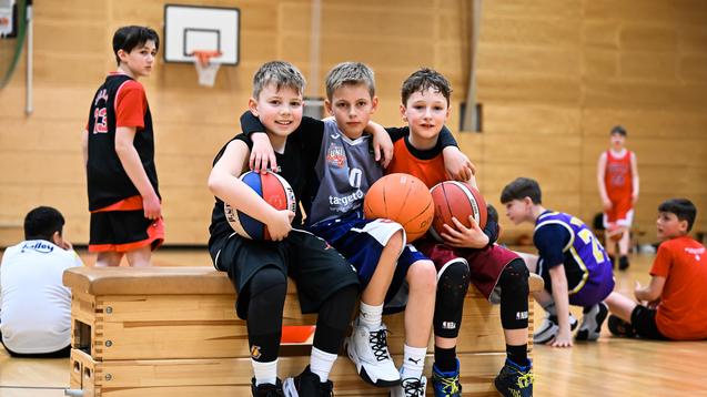 Drei Kinder posieren auf einer Bank in einer Sporthalle, jeder hält einen Basketball. Im Hintergrund weitere Spieler.