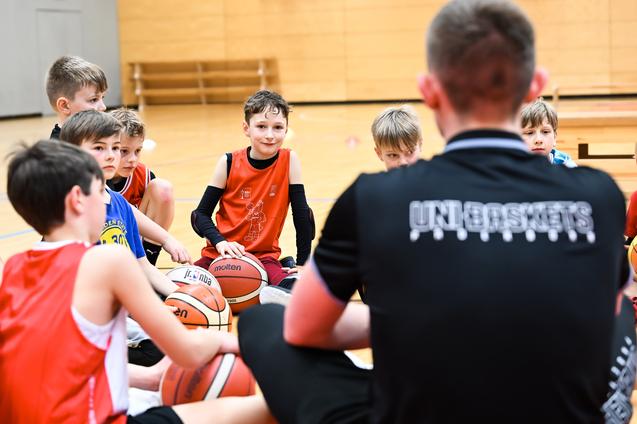 Gruppe von Jungen beim Basketballtraining, sitzend auf dem Boden mit Bällen, Trainer spricht zu ihnen.