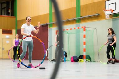 Eine Frau hält einen bunten Hula-Hoop-Reifen und lächelt, während zwei weitere Frauen im Hintergrund üben.