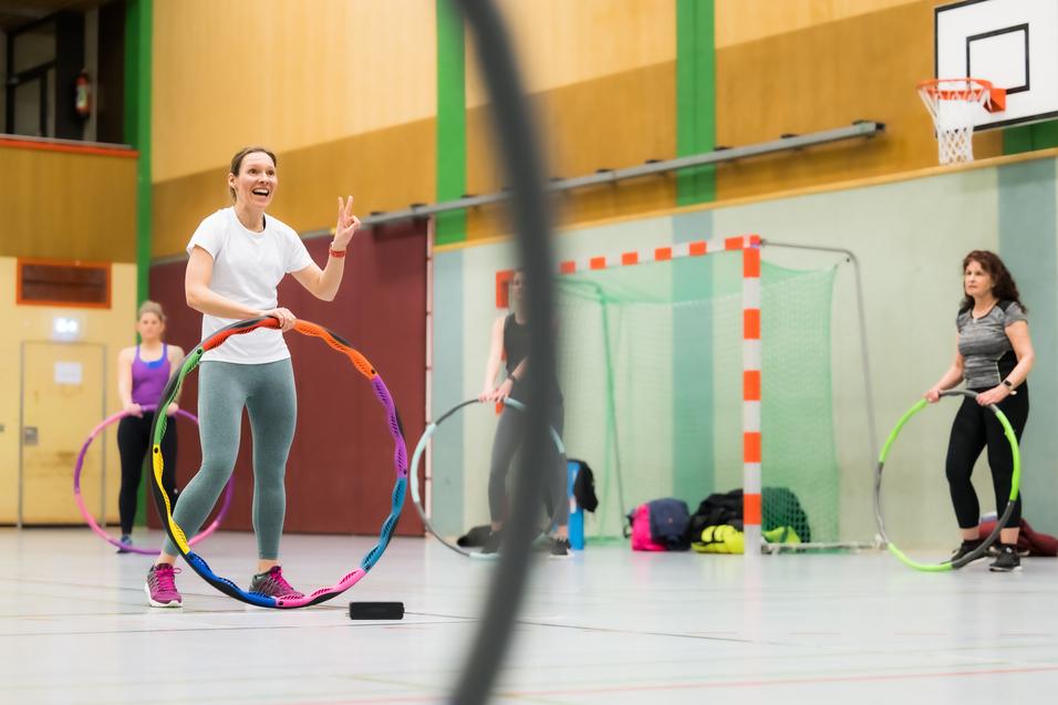 Eine Frau hält einen bunten Hula-Hoop-Reifen und lächelt, während zwei weitere Frauen im Hintergrund üben.