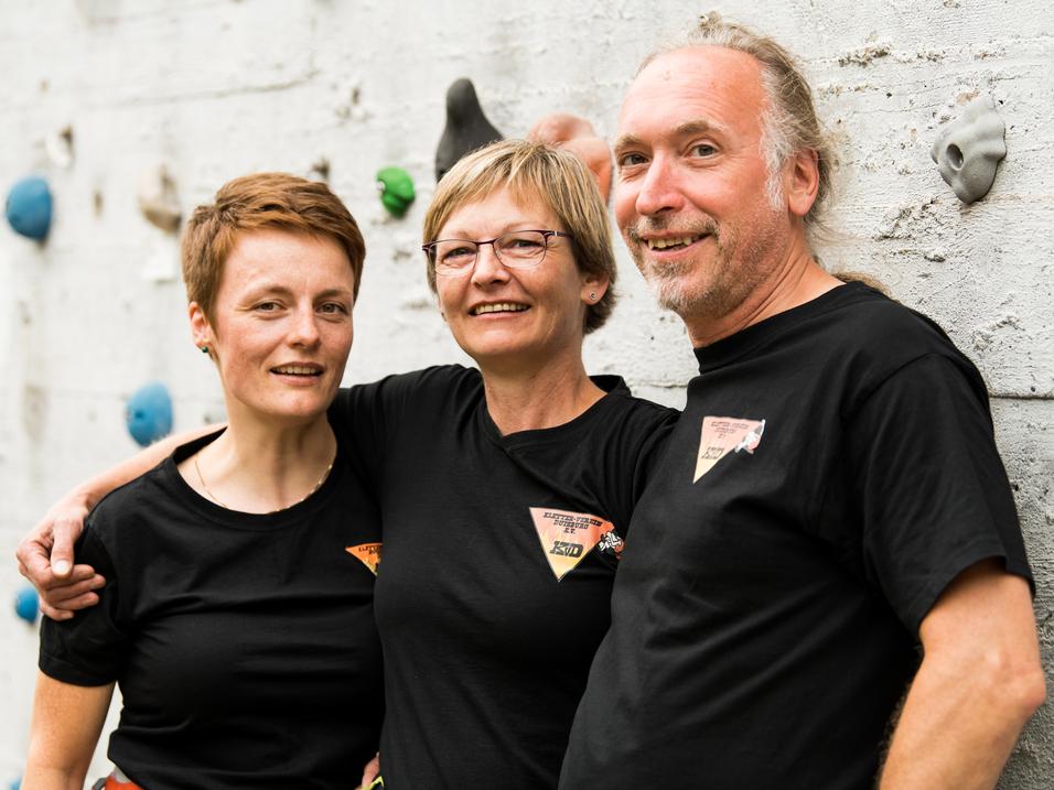 Drei Personen in schwarzen T-Shirts lächeln vor einer Kletterwand mit bunten Griffen.