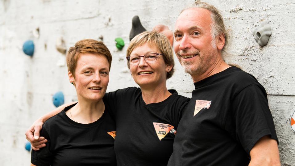 Drei Personen in schwarzen T-Shirts lächeln vor einer Kletterwand mit bunten Griffen.