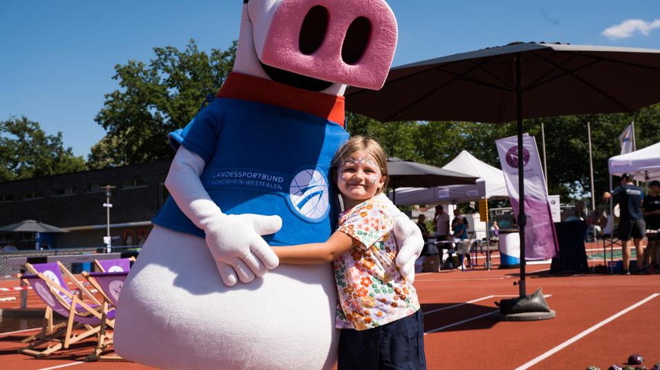 Ein Kind umarmt ein Maskottchen in Sportbekleidung auf einem Sportplatz mit Ständen und Sportequipment im Hintergrund.