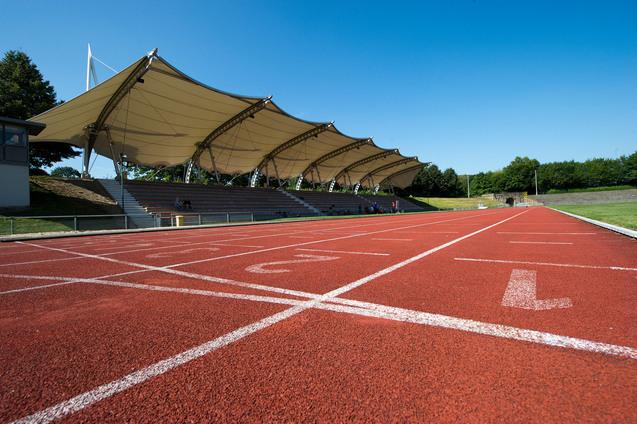Nahaufnahme einer roten Laufbahn mit Startlinien und einem überdachten Zuschauerbereich im Hintergrund.