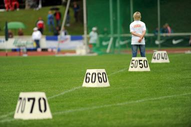 Person mit kurzen Haaren steht auf einem Sportplatz vor nummerierten Markierungen (40, 50, 60, 70) im Gras.