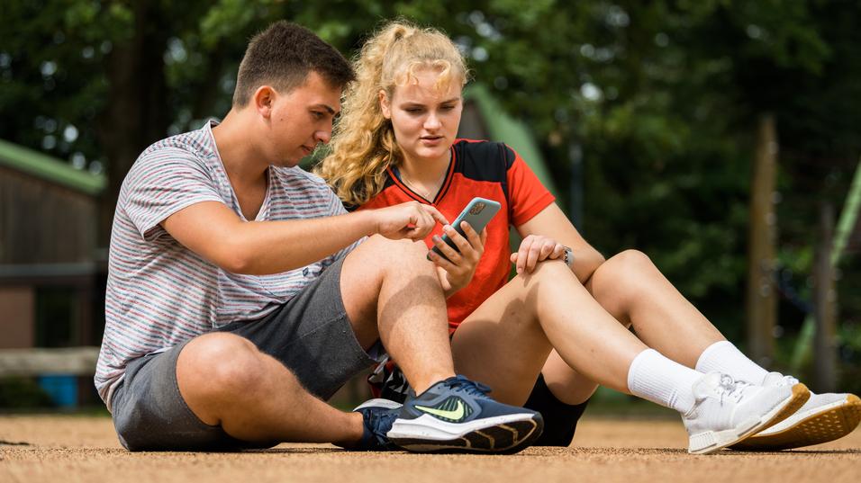 Zwei Jugendliche sitzen auf dem Boden und betrachten gemeinsam ein Smartphone. Ein Mädchen trägt ein rotes sportliches Oberteil.