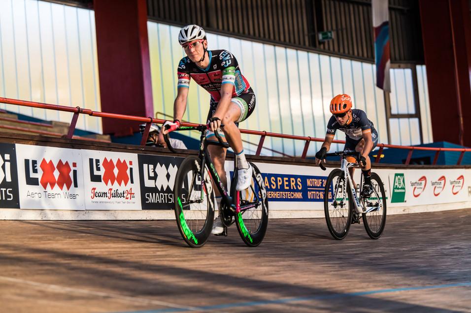Zwei Radrennfahrer auf einer Holzbahn, einer mit weiß-emblemierter Ausrüstung und der andere mit orangefarbener Helm.