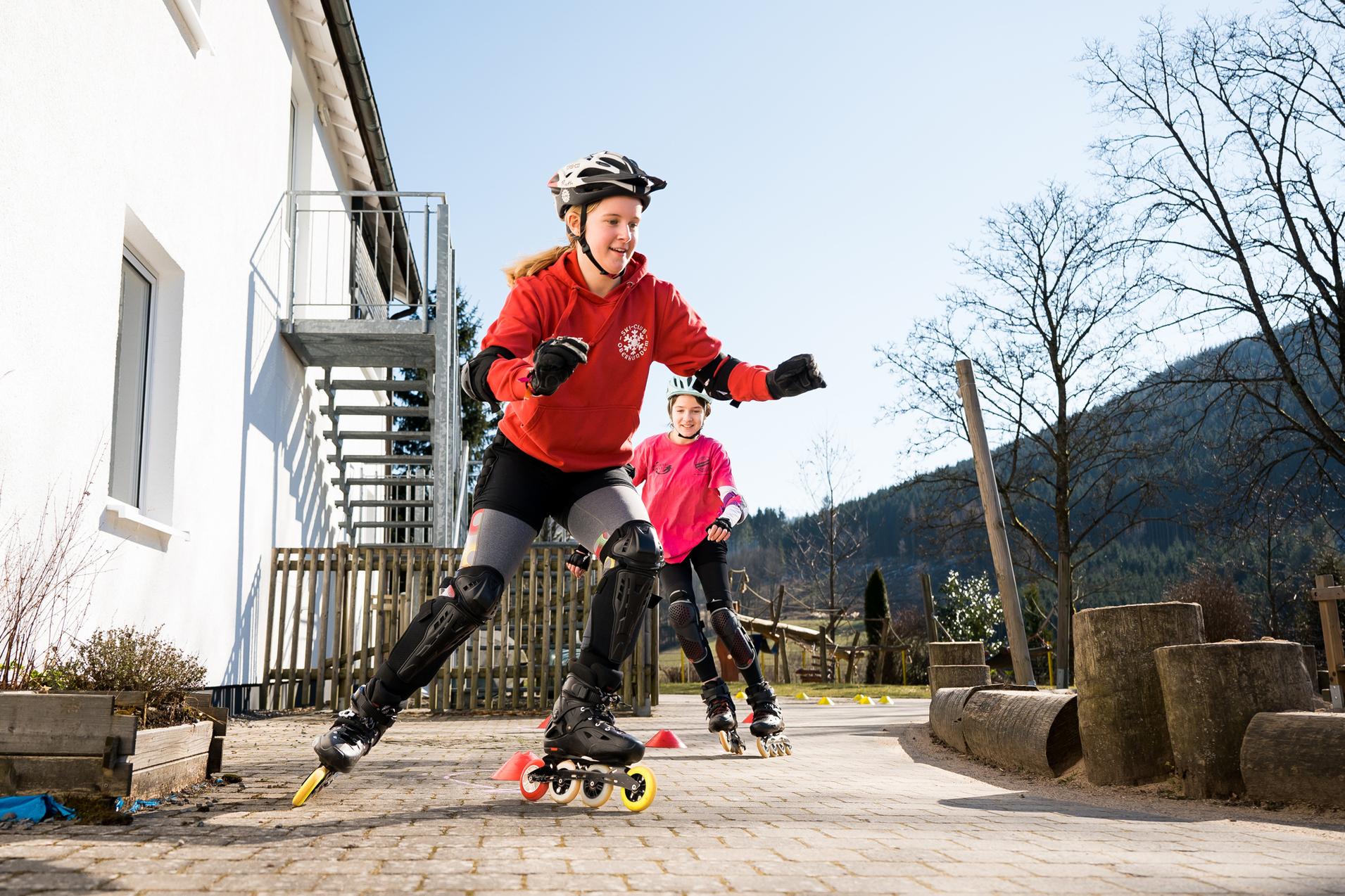 Junge Skaterinnen fahren auf Rollschuhen auf einem gepflasterten Weg, umgeben von Bäumen und einem weißen Gebäude.