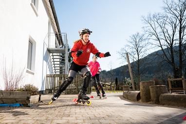 Junge Skaterinnen fahren auf Rollschuhen auf einem gepflasterten Weg, umgeben von Bäumen und einem weißen Gebäude.
