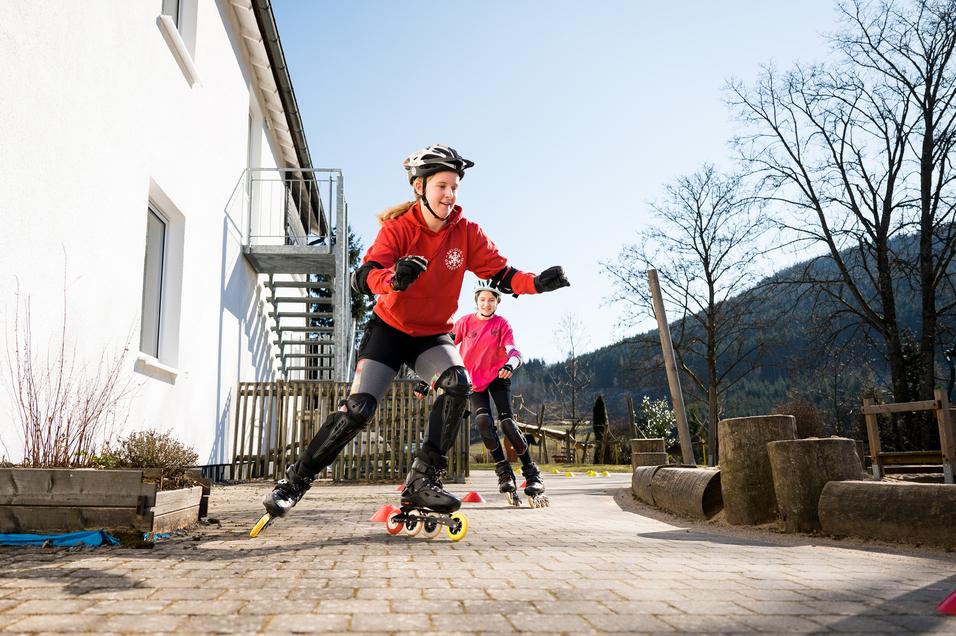 Junge Skaterinnen fahren auf Rollschuhen auf einem gepflasterten Weg, umgeben von Bäumen und einem weißen Gebäude.