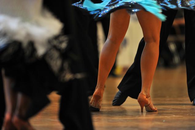Tanzende Beine in eleganten Schuhen, umgeben von schwingenden Kostümen auf einem Holzboden.