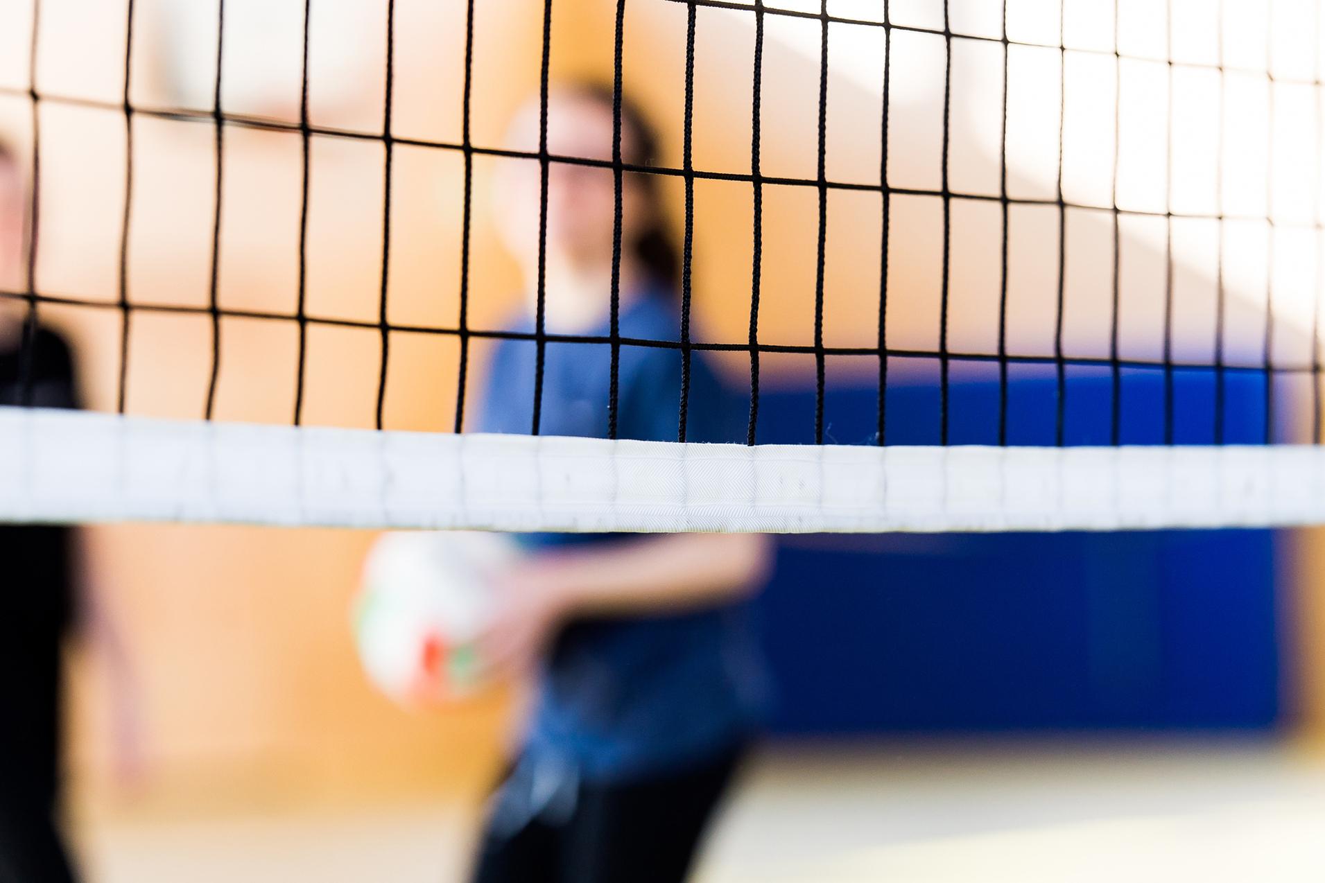 Unscharfe Silhouetten von zwei Personen hinter einem Volleyballnetz in einer Sporthalle. Eine Person hält einen Volleyball.