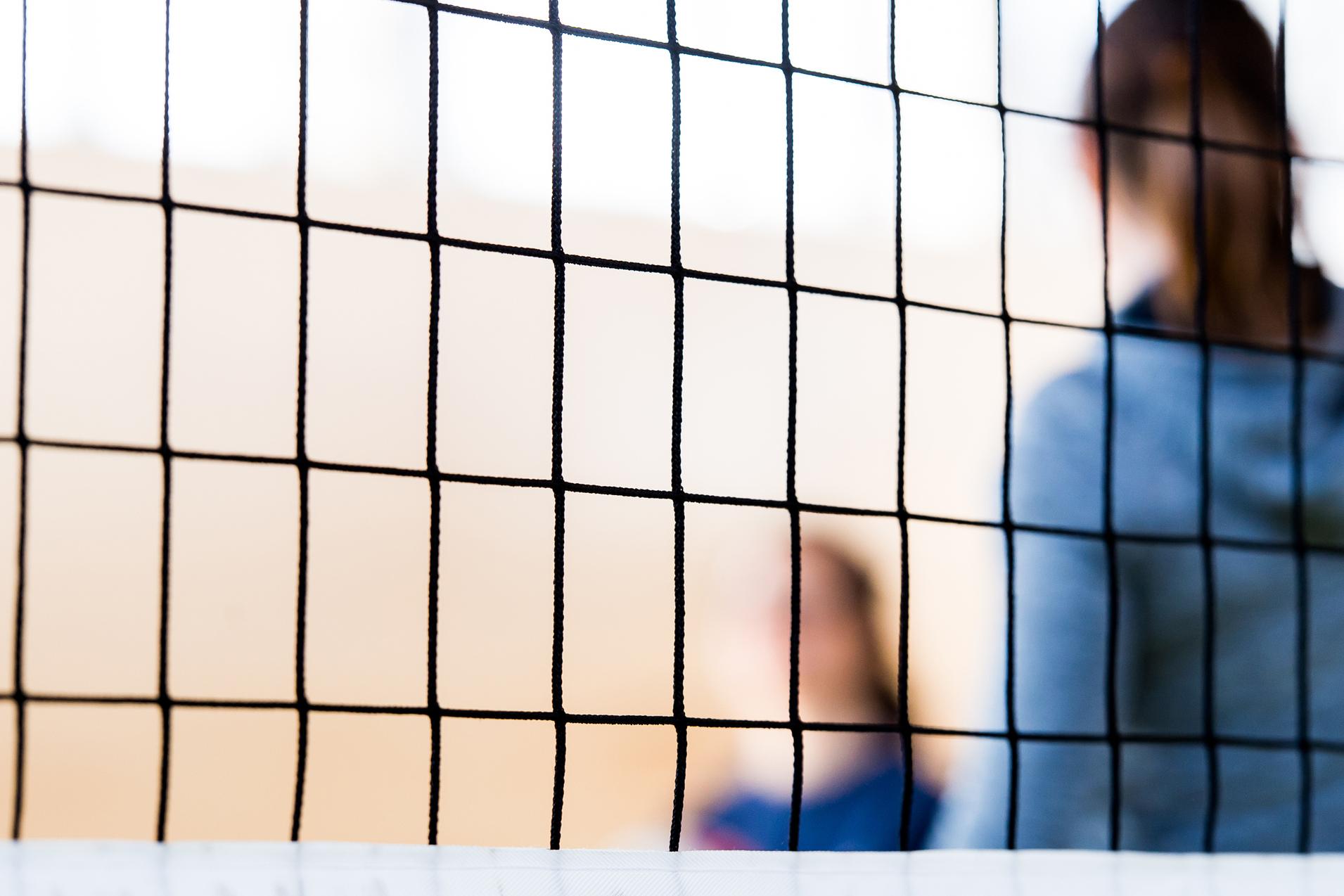 Nahaufnahme eines Volleyballnetzes mit unscharfen Spielern im Hintergrund in einer Sporthalle.